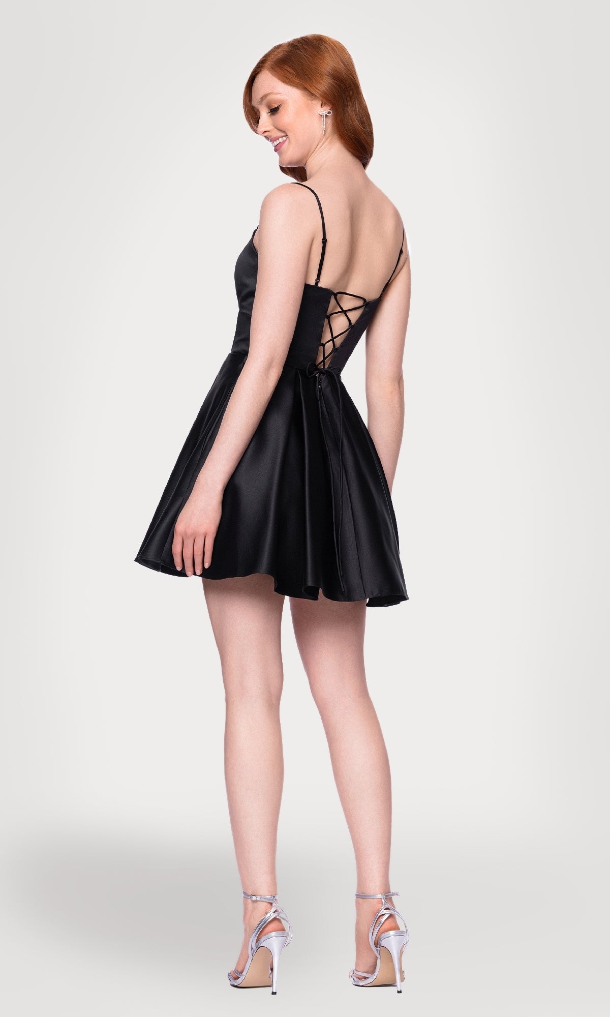 A woman models the Cut-Out Black Mini Hoco Dress: Blondie Nites 5605BN, featuring a short black satin design with a lace-up back. She stands in silver heels against a plain white background, facing slightly away from the camera.